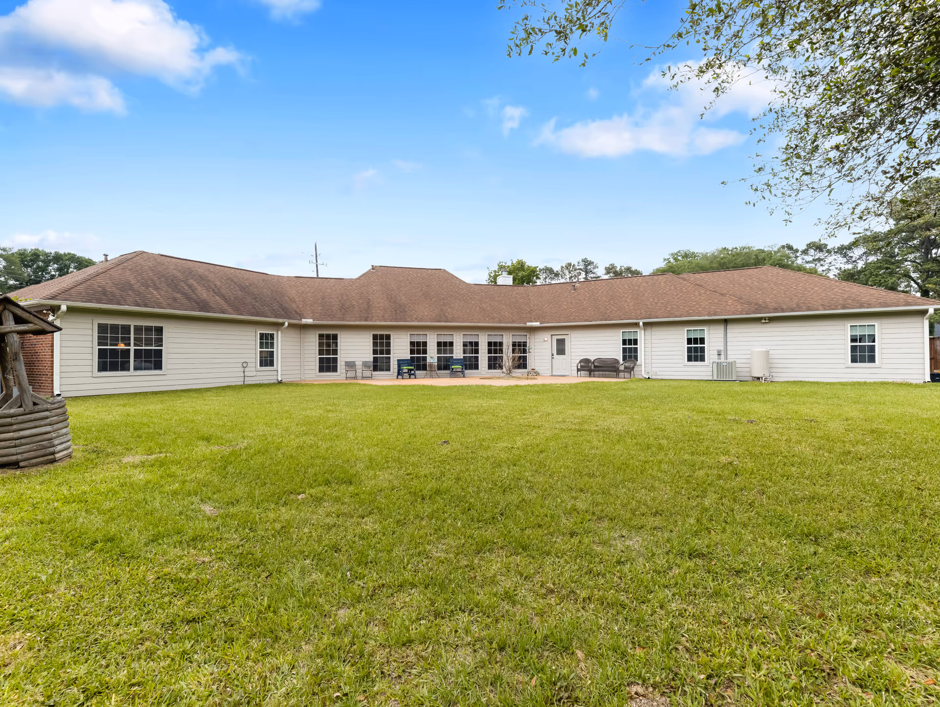 Wide exterior view of a single-story building with a brown roof and white siding, surrounded by a large green lawn under a blue sky with some clouds. There are several windows and a door facing the lawn, with outdoor seating on a small patio area. Trees are visible on the right side.