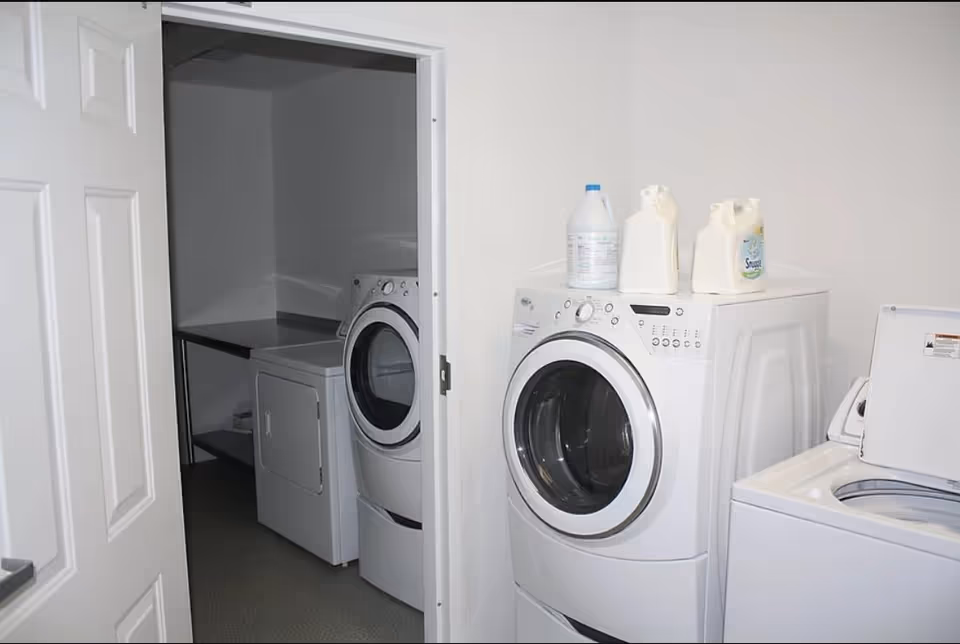 Laundry room with a front-loading washing machine and dryer, a top-loading washing machine, and laundry detergent bottles placed on top of the front-loading washing machine. The room has white walls and a door partially open to reveal another dryer and a countertop inside.