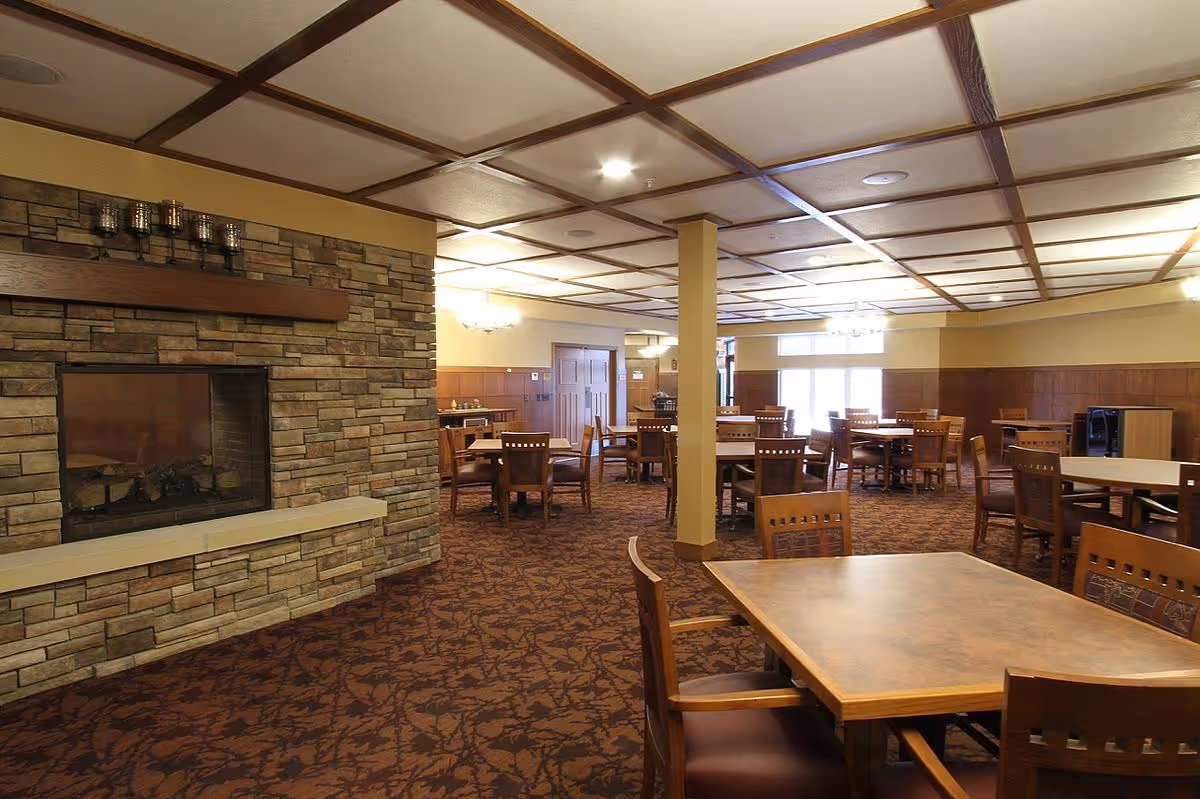 Spacious dining room with multiple wooden tables and chairs arranged neatly. The room features a stone fireplace on the left side and a coffered ceiling with recessed lighting. The carpet has a dark pattern, and the walls are decorated with wood paneling and light-colored paint. Large windows at the far end allow natural light to enter the room.