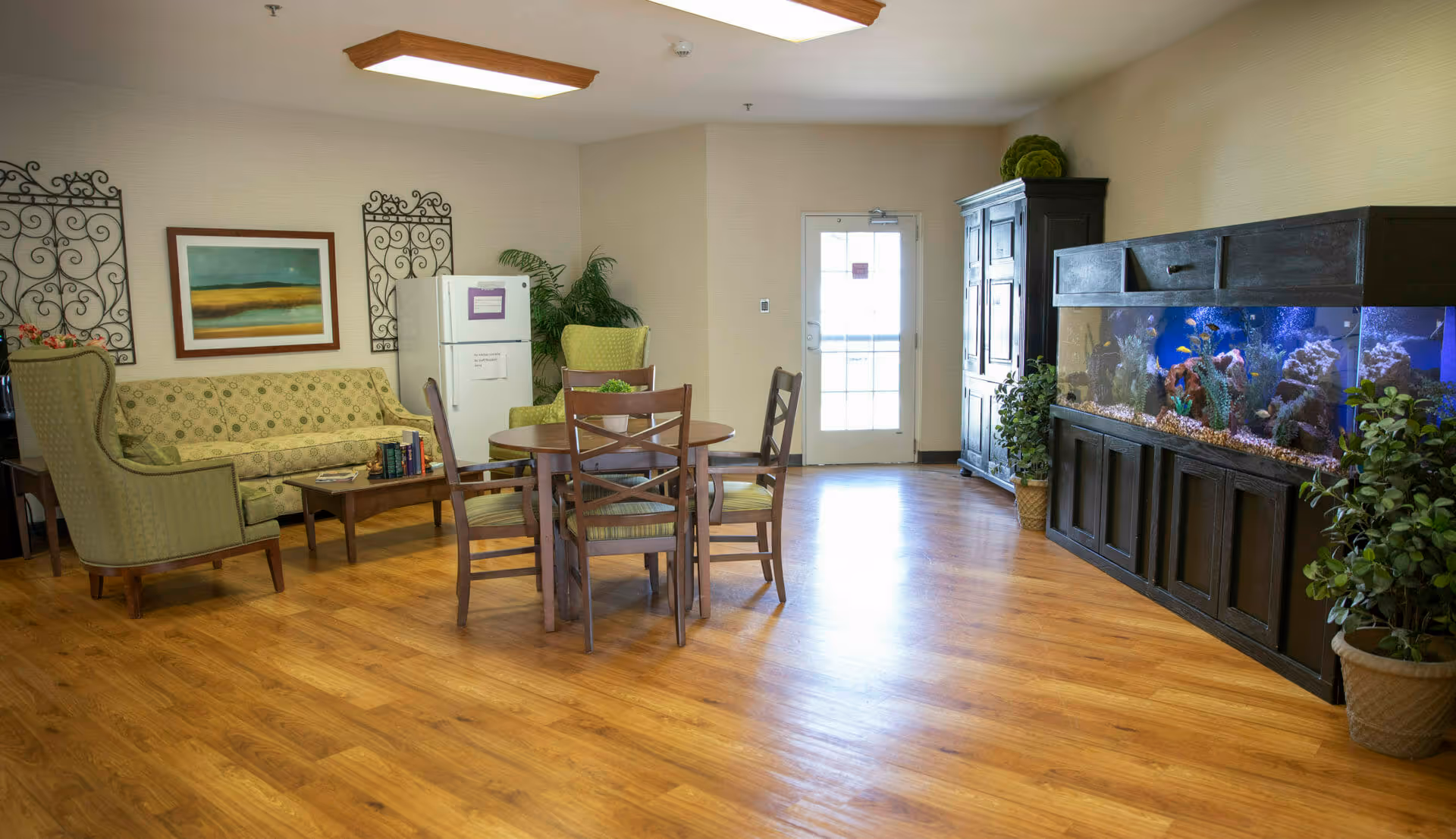 A cozy common area in Brownsburg Meadows featuring a round wooden table with four chairs, a green patterned sofa, two green armchairs, a large aquarium with colorful fish, potted plants, a white refrigerator, and a door with a window letting in natural light.