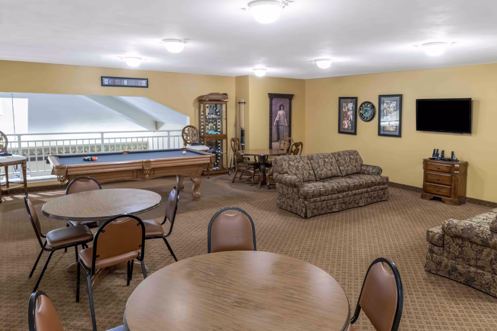 Community lounge with a pool table, round tables and chairs, upholstered sofas, and a wall-mounted TV.