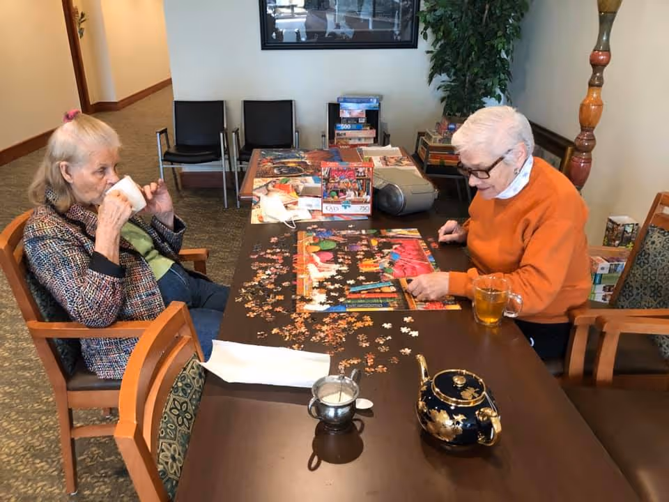Two elderly women sitting at a table in a common area, one drinking from a cup and the other working on a colorful jigsaw puzzle. The table has a teapot, a cup of tea, and puzzle pieces scattered around. There are chairs, a plant, and board games in the background.