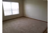Empty room with beige carpet, white walls, a large window with blinds, and wooden baseboards.