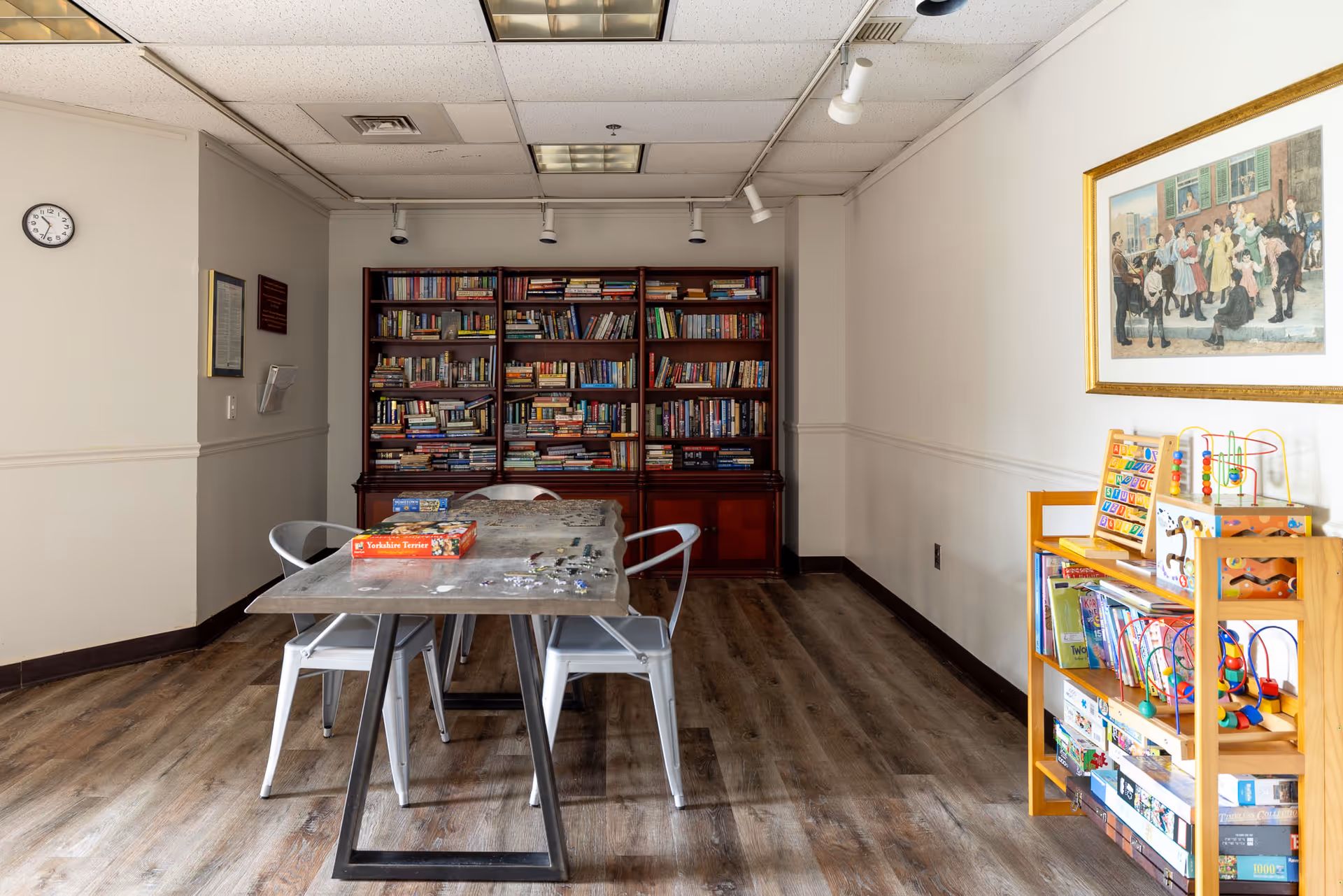 A room with a wooden table and four metal chairs, a large bookshelf filled with books against the back wall, and a wooden shelf on the right holding children's toys and board games. A framed painting hangs on the right wall, and a clock is mounted on the left wall above a small plaque.