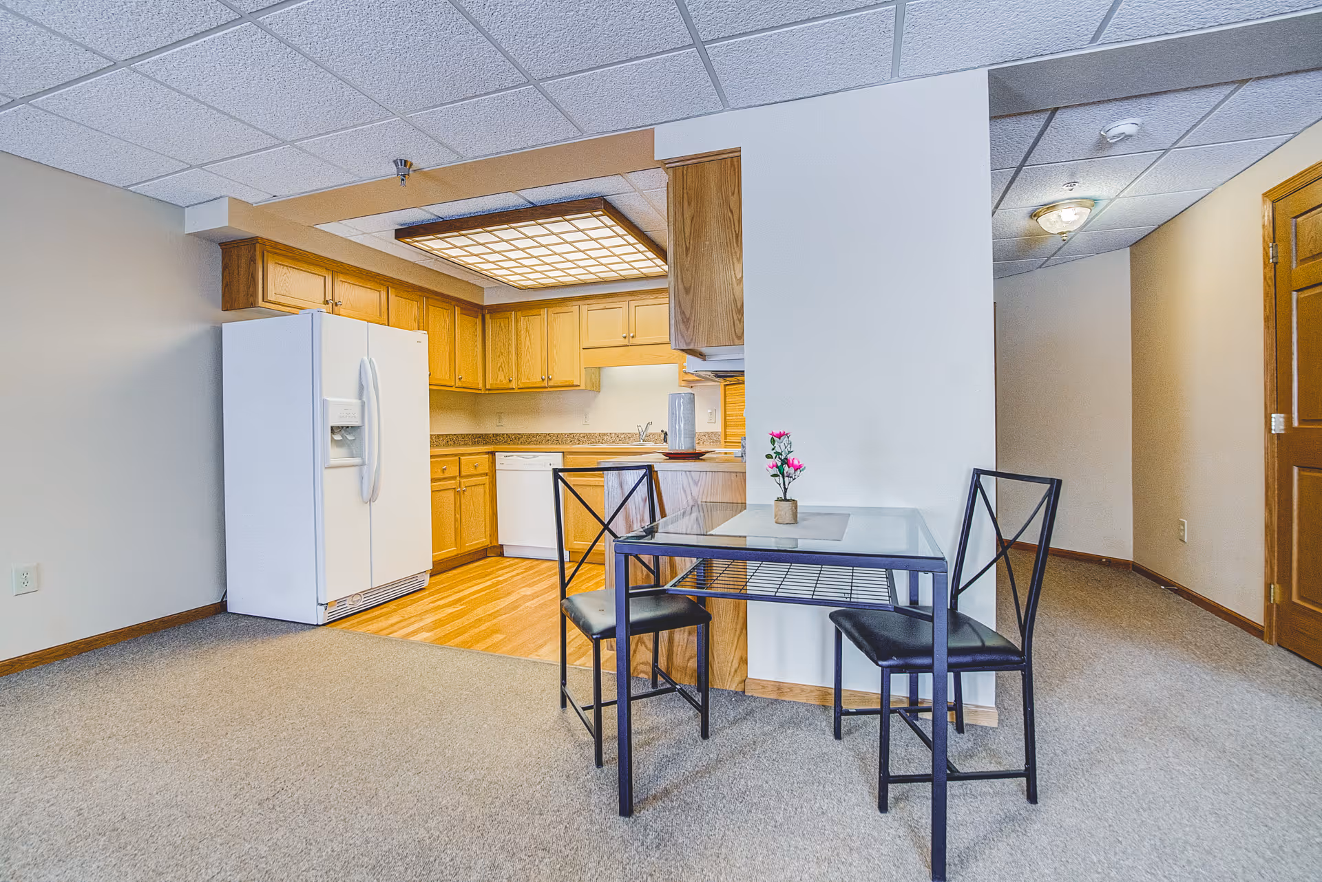 Open kitchenette with oak cabinets and a white refrigerator next to a small glass dining table with two chairs on a carpeted floor.