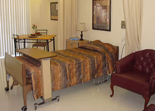 A single hospital-style bed with a brown patterned blanket in a room. Next to the bed is a small wooden nightstand with a lamp and a framed picture on the wall above it. There is a small table with two chairs near a window with vertical blinds, and a maroon upholstered armchair is positioned near the bed.