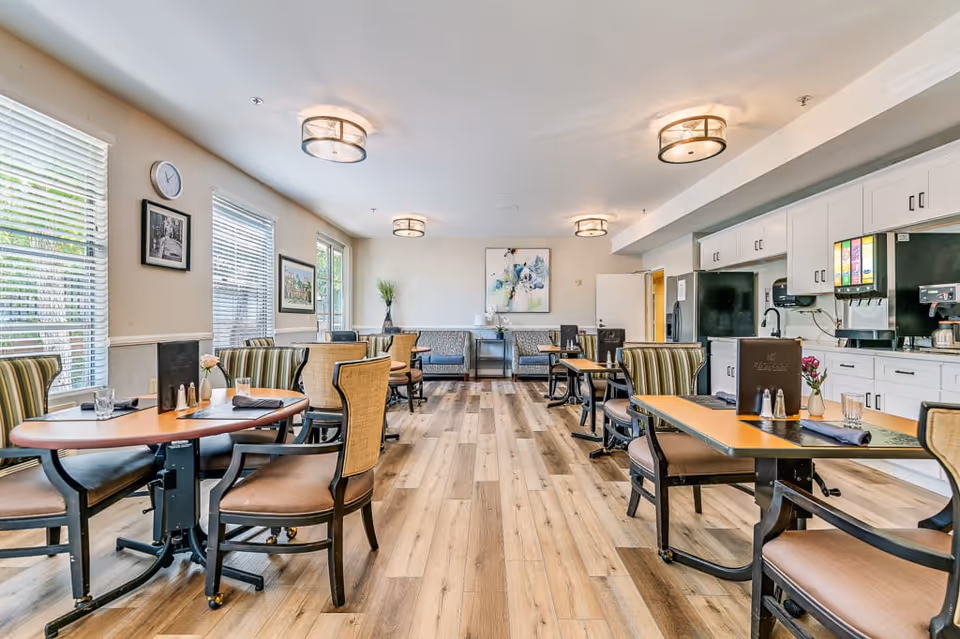 A bright and spacious dining room in a senior living facility with wooden floors, multiple tables and chairs arranged neatly, large windows with blinds letting in natural light, white cabinets along one wall with beverage dispensers, and modern ceiling lights.