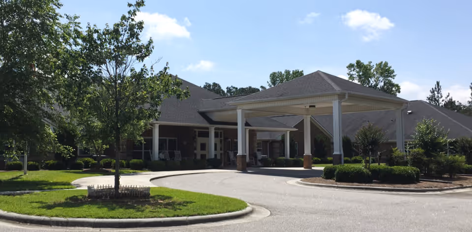Exterior view of Prestwick Village senior living facility showing a covered entrance with white pillars, surrounded by green trees and shrubs under a blue sky with some clouds.