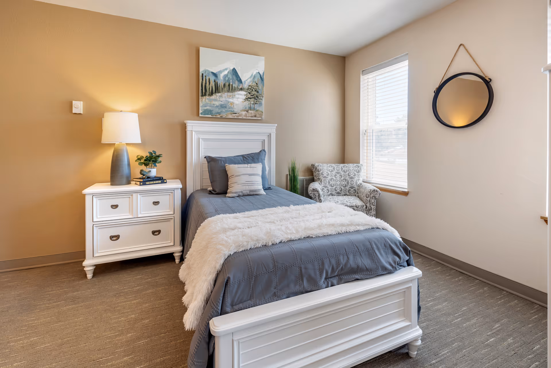 Cozy bedroom with a white single bed dressed in gray bedding, a white nightstand with a lamp, a patterned armchair, and a window.