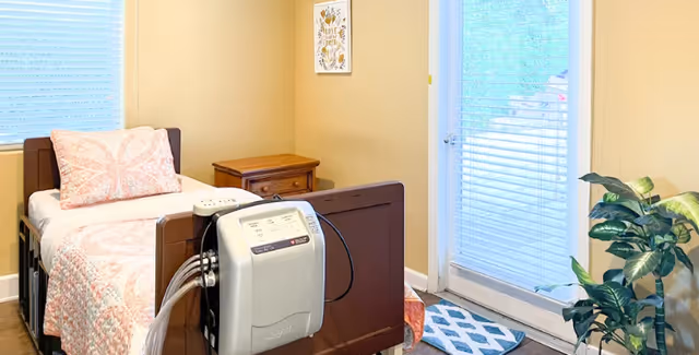 A well-lit bedroom in a senior living facility featuring a hospital-style bed with a pink and white quilt and pillow. Next to the bed is a wooden nightstand with a drawer. A medical device is attached to the foot of the bed. The room has beige walls, a window with blinds, a glass door with blinds leading outside, a small rug, and a green potted plant in the corner.