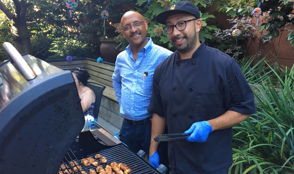 Two men standing outdoors near a barbecue grill with cooked chicken pieces on it. One man is wearing a blue checkered shirt and glasses, smiling at the camera. The other man is wearing a black chef's coat, a black cap, glasses, and blue gloves, holding tongs and smiling. There are green plants and decorative hanging ornaments in the background.