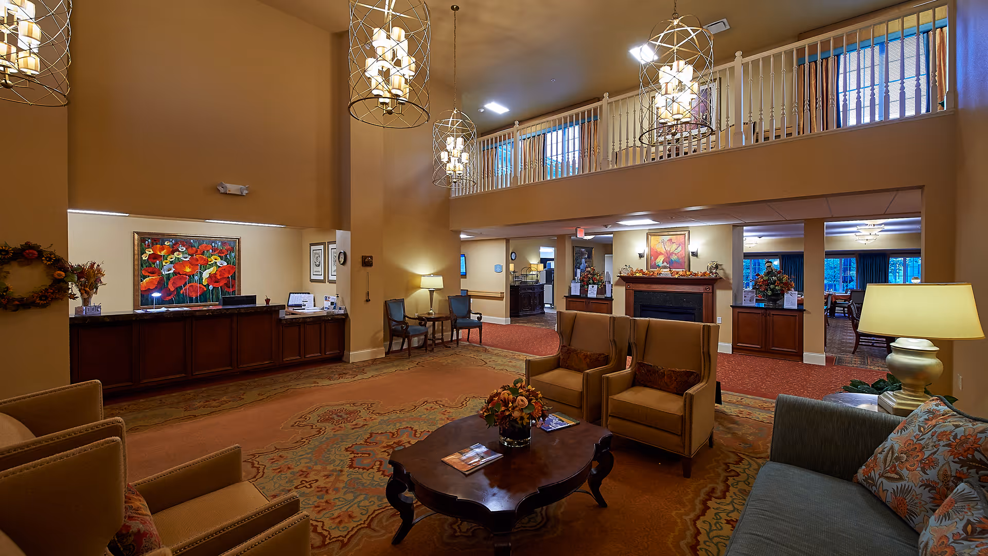 A spacious and warmly lit senior living facility lobby with high ceilings and hanging pendant lights. The area features comfortable seating including armchairs and a sofa around a wooden coffee table with a floral centerpiece. There is a reception desk with a colorful floral painting behind it, a fireplace with autumn-themed decorations, and a balcony railing overlooking the lobby. The decor includes warm tones, patterned carpets, and framed artwork on the walls.