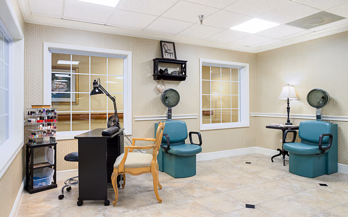 A beauty salon area inside a senior living facility featuring two green salon chairs with hair dryers, a black manicure table with a lamp, a wooden chair, a small table with a lamp, and a rack with various nail polish bottles. The room has beige patterned wallpaper, tiled floor, and two windows with white frames.