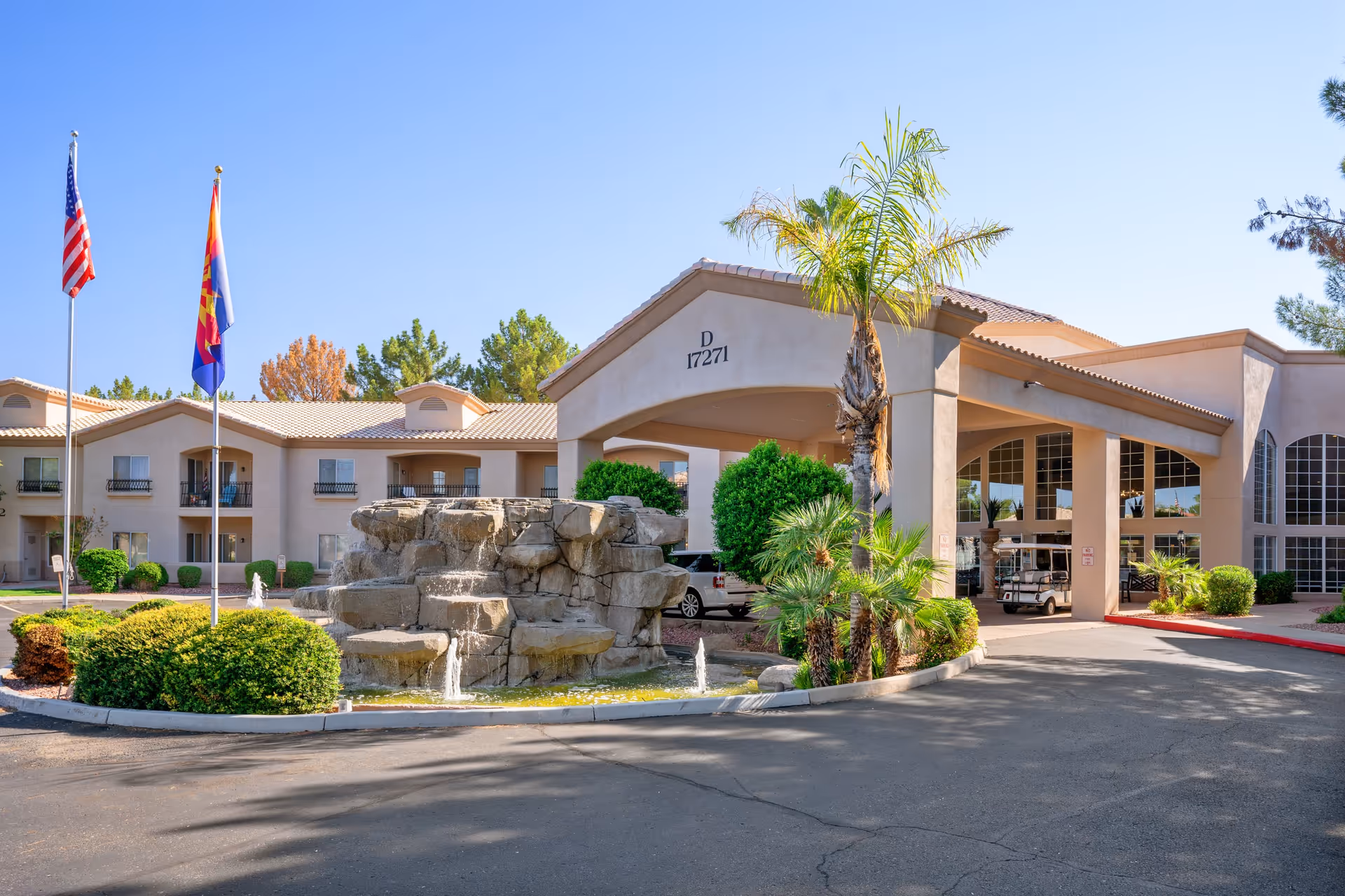 Exterior view of The Montecito Senior Living facility featuring a beige stucco building with a covered entrance, a rock water fountain in front, two flagpoles with the American and Arizona state flags, and surrounding greenery including palm trees and bushes under a clear blue sky.