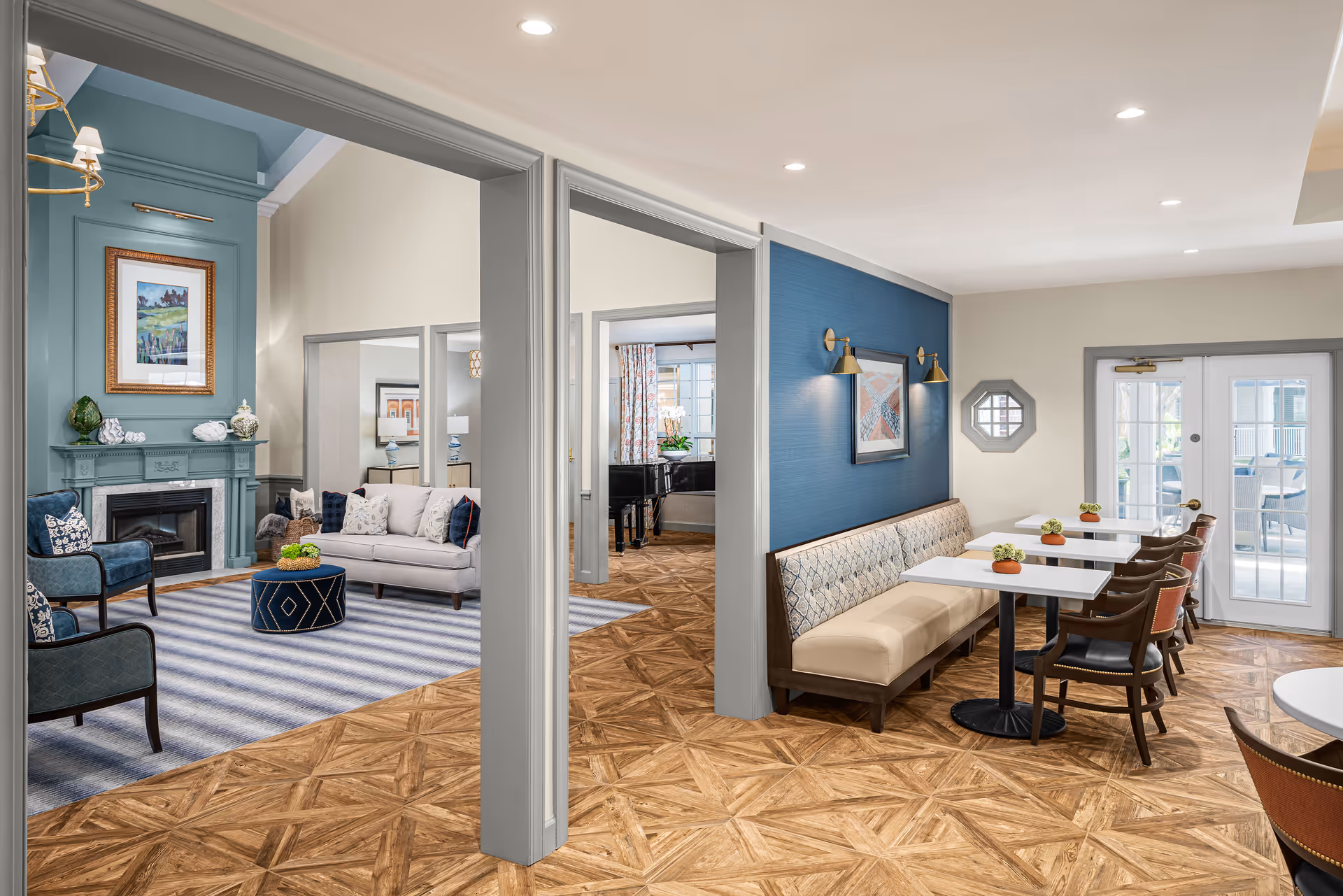 A bright and spacious interior common area in a senior living facility featuring a seating area with a fireplace, armchairs, and a sofa on a striped rug. Adjacent to it is a dining area with tables, chairs, and a cushioned bench against a blue accent wall with wall-mounted lights and artwork. The room has wooden patterned flooring and large windows allowing natural light.