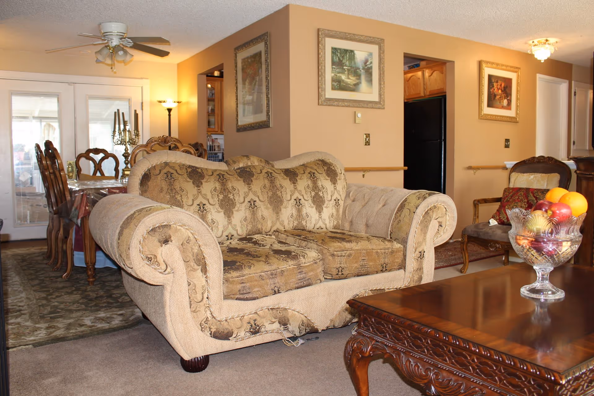 A cozy living room area with a patterned beige loveseat and a wooden coffee table holding a glass bowl filled with fruit. In the background, there is a dining area with a wooden dining table and chairs, a ceiling fan with lights, framed paintings on the walls, and a glimpse of a kitchen with a black refrigerator.