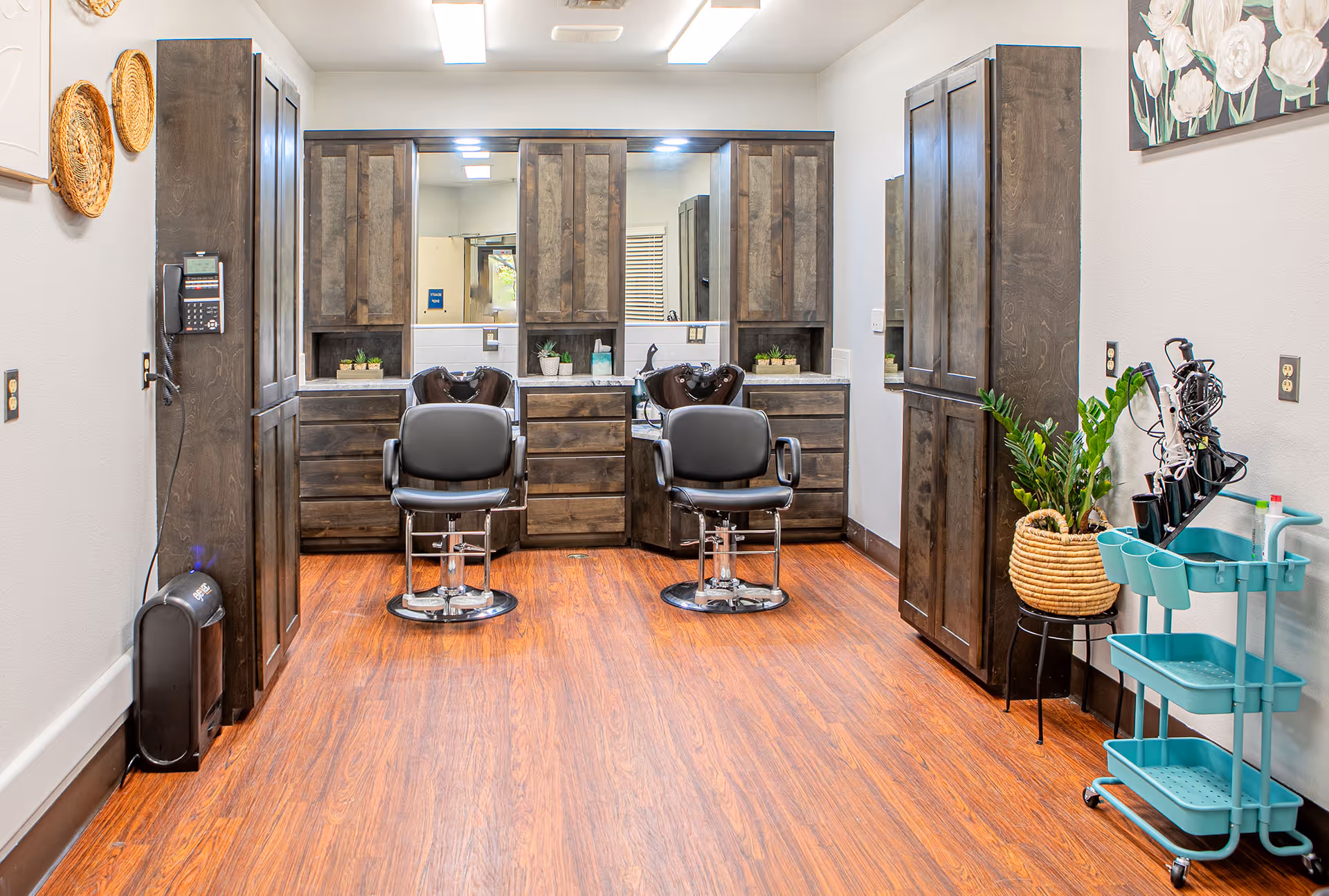 Interior view of a salon area in a senior living facility with two black salon chairs in front of large mirrors and wooden cabinets. The room has wood flooring, a plant in a woven basket on a stand, a turquoise rolling cart with hair styling tools, and decorative wall hangings.