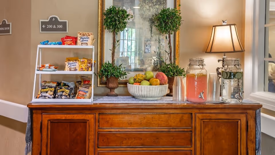 A wooden sideboard with a white snack rack holding various packaged snacks, a large bowl of assorted fresh fruit, two glass beverage dispensers filled with pink lemonade and water with cucumber slices, a table lamp with a beige shade, and two potted green plants. A large framed mirror hangs on the wall behind the sideboard, reflecting a window and part of the room.