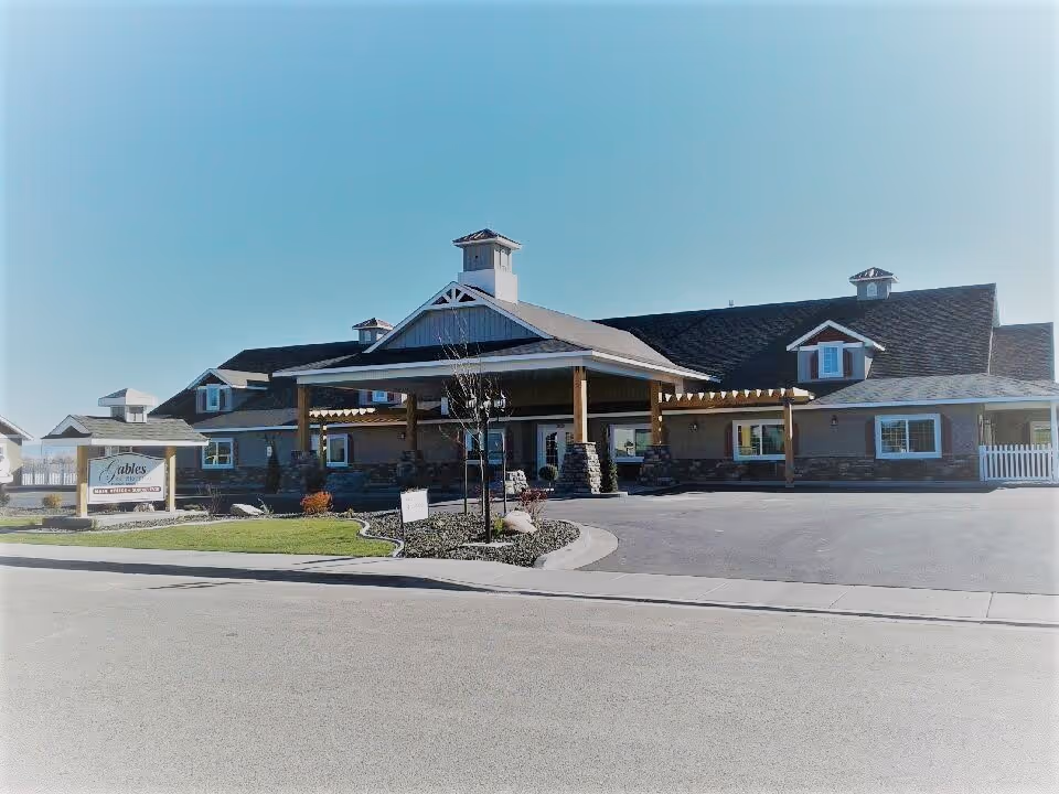 Front exterior of The Gables Assisted Living building showing a covered porte-cochère, landscaped driveway island, and an entrance sign.