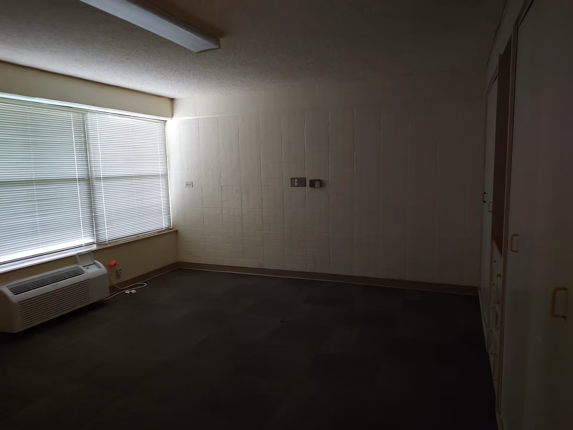Empty room with dark carpet flooring, a large window with closed blinds on the left wall, a wall-mounted air conditioning unit below the window, and built-in cabinets on the right side. The walls are light-colored with a textured pattern, and there are two wall switches or controls in the center of the far wall.