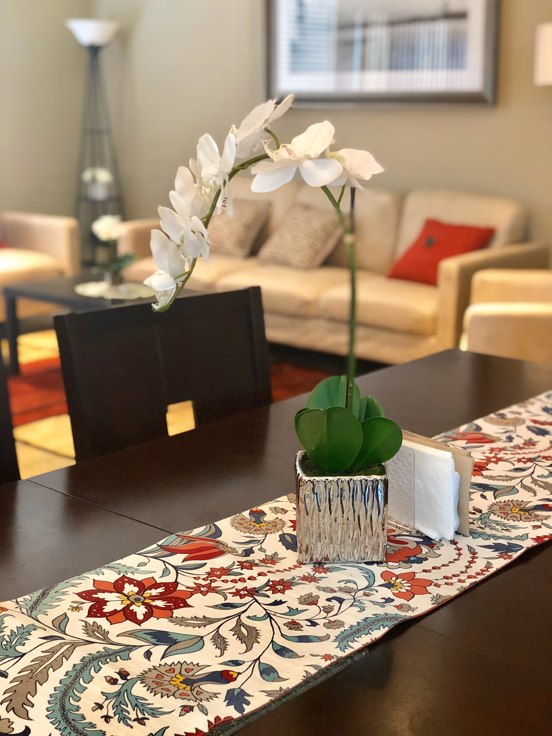 A close-up view of a dark wooden dining table with a colorful floral table runner. On the table is a silver vase holding a white orchid plant and a napkin holder. In the background, there is a beige sofa with patterned and red cushions, a floor lamp, and a framed picture on the wall.