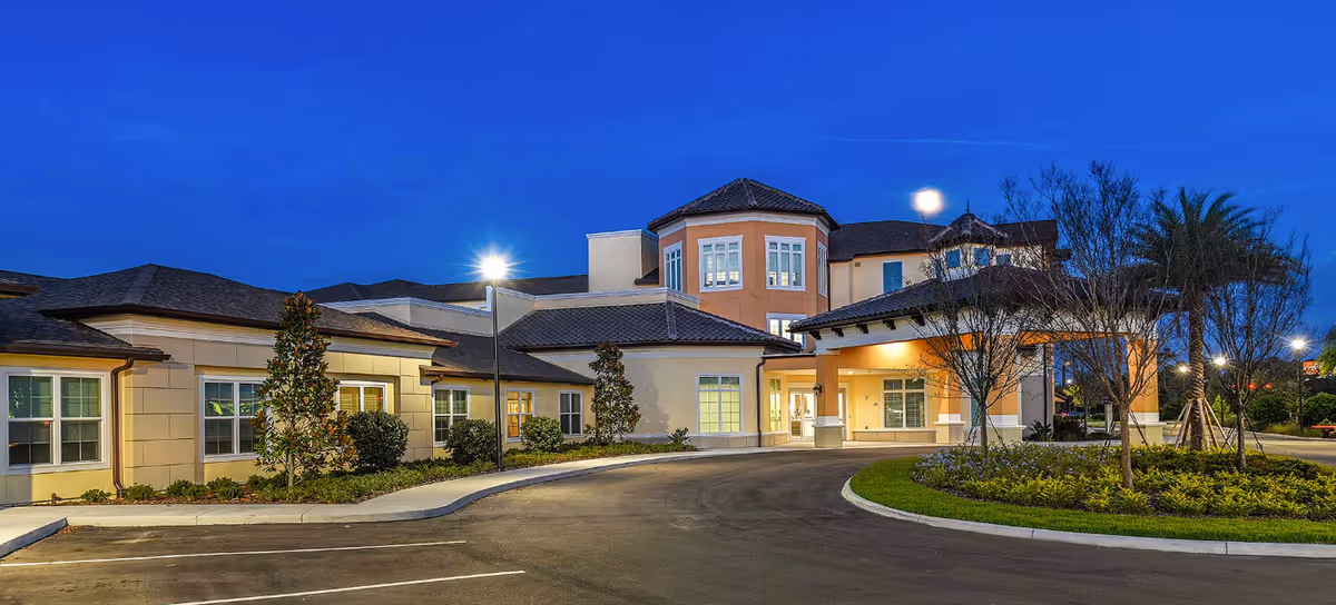 Well-lit modern senior living facility entrance with a circular driveway, landscaping, and palm trees at dusk.