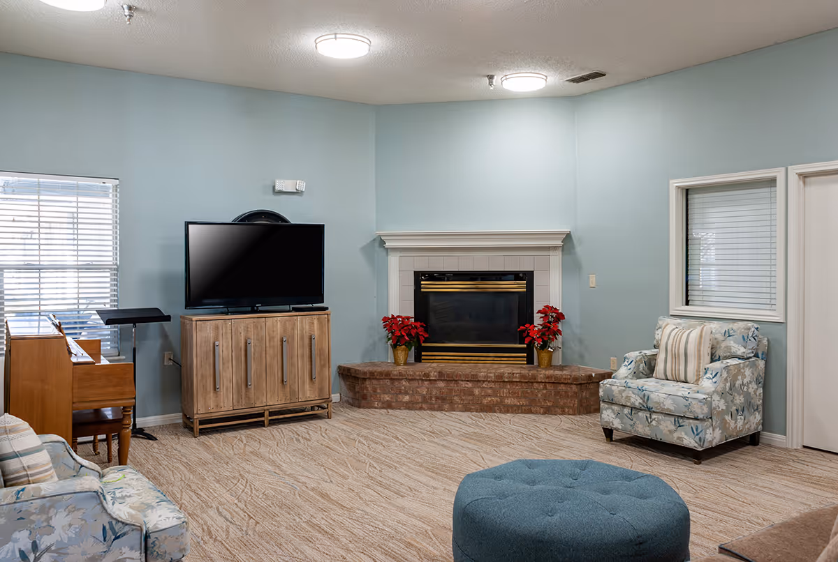 A cozy living room with light blue walls, a brick fireplace decorated with two red poinsettia plants, a flat-screen TV on a wooden cabinet, a floral upholstered armchair with a striped pillow, a piano with a music stand, and a round blue ottoman on a beige carpeted floor.