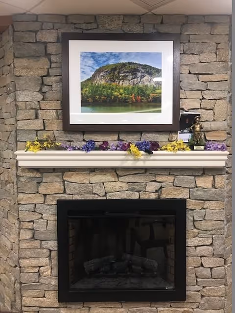 Stone fireplace with a black framed glass front. Above the fireplace is a white mantel decorated with yellow and purple flowers. A framed photograph of a rocky hill with trees and a body of water is hung on the stone wall above the mantel.