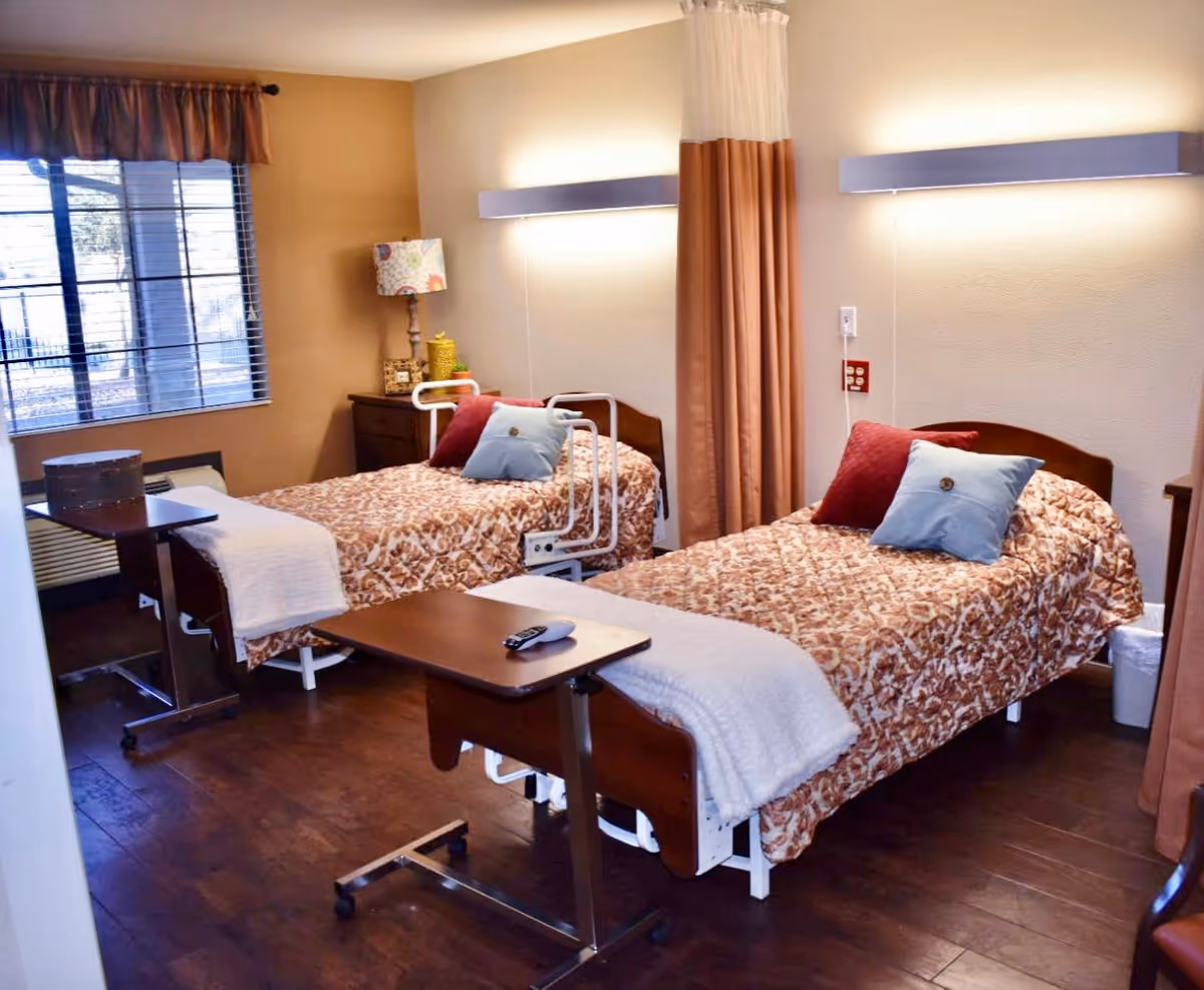A nursing facility bedroom with two single beds, each covered with a patterned bedspread and adorned with two pillows. Each bed has a bedside table with a lamp and a rolling overbed table in front. The room has wooden flooring, a window with blinds and a valance, and a privacy curtain between the beds.