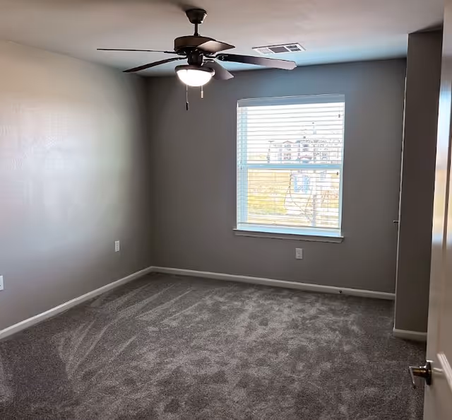 Empty, gray-carpeted interior room with a ceiling fan and window blinds.