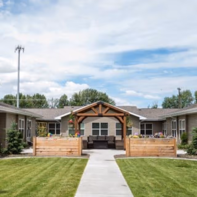 Outdoor courtyard area of The Gables Assisted Living of Idaho Falls featuring a wooden pergola with seating underneath, surrounded by raised wooden planter boxes with flowers, green grass, and a concrete walkway leading to the seating area. The building exterior is visible in the background under a partly cloudy sky.