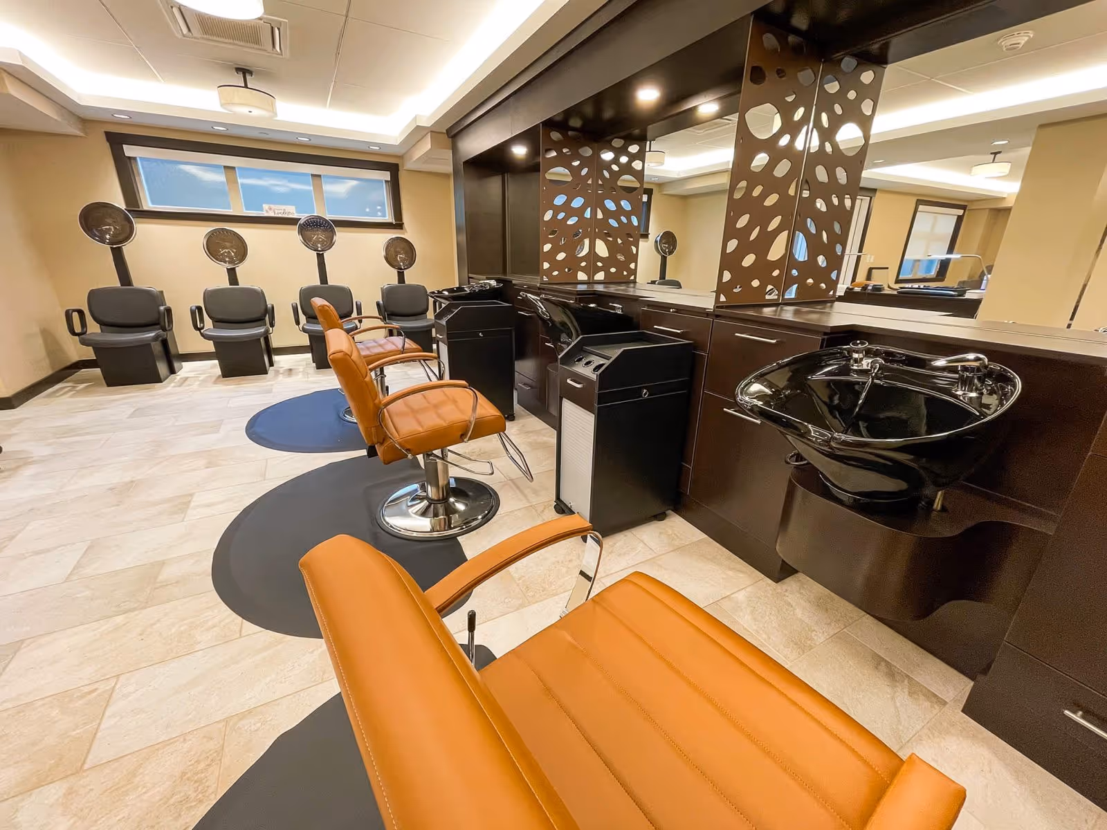 Interior view of a hair salon area with three brown leather styling chairs facing mirrors and black wash basins. In the background, there are four black hair drying chairs with hooded dryers. The room has beige walls, a tiled floor, and a window near the ceiling.