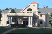 Front exterior view of a two-story nursing and rehabilitation center building with a covered entrance supported by white columns, surrounded by greenery and a clear sky.