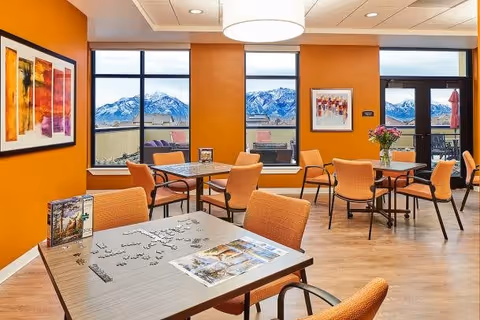 A bright and spacious common area with orange walls and large windows showcasing a mountain view. The room contains several tables with orange chairs, one table has a partially completed puzzle and a brochure. There is a door leading to an outdoor patio with seating and umbrellas. Artwork is displayed on the walls and a ceiling light fixture is visible.