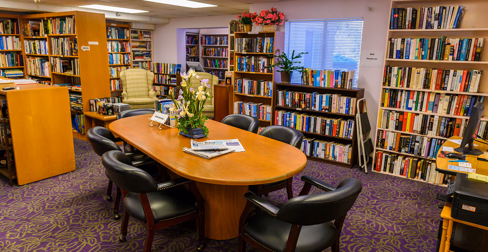 A cozy library/reading room with a wooden oval table surrounded by black chairs, bookshelves lining the walls, and a purple patterned carpet.