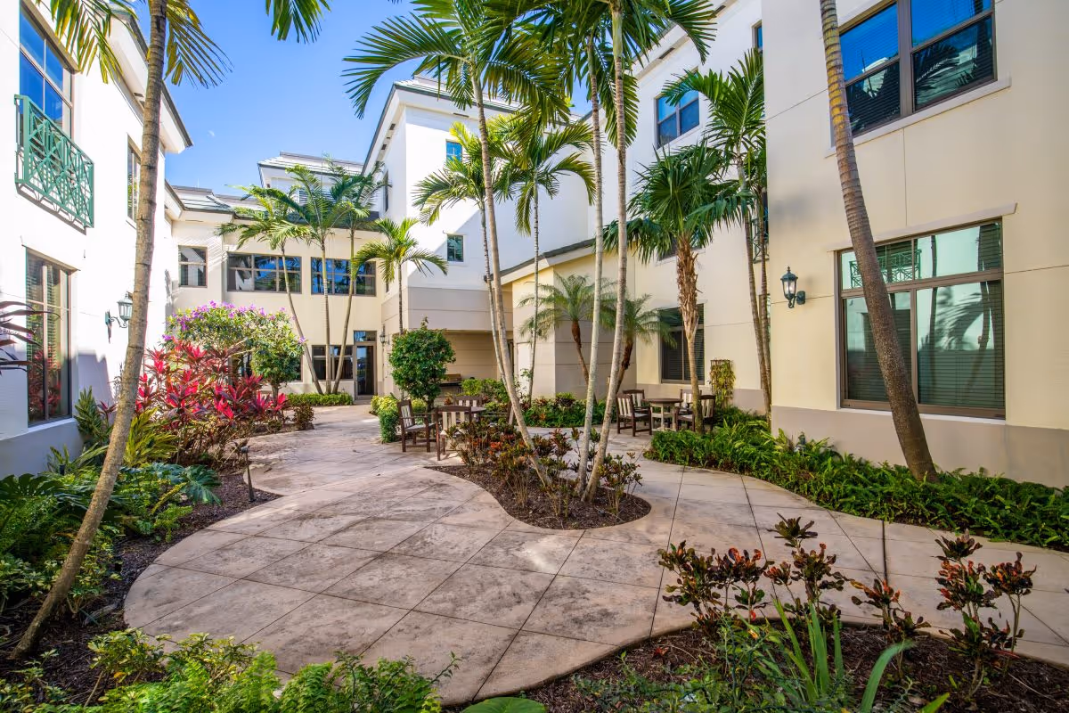 Outdoor courtyard area at Luxe Senior Living at Wellington featuring a paved walkway surrounded by tropical plants and palm trees, with several wooden chairs and tables arranged for seating. The courtyard is enclosed by a multi-story building with large windows and light-colored walls.
