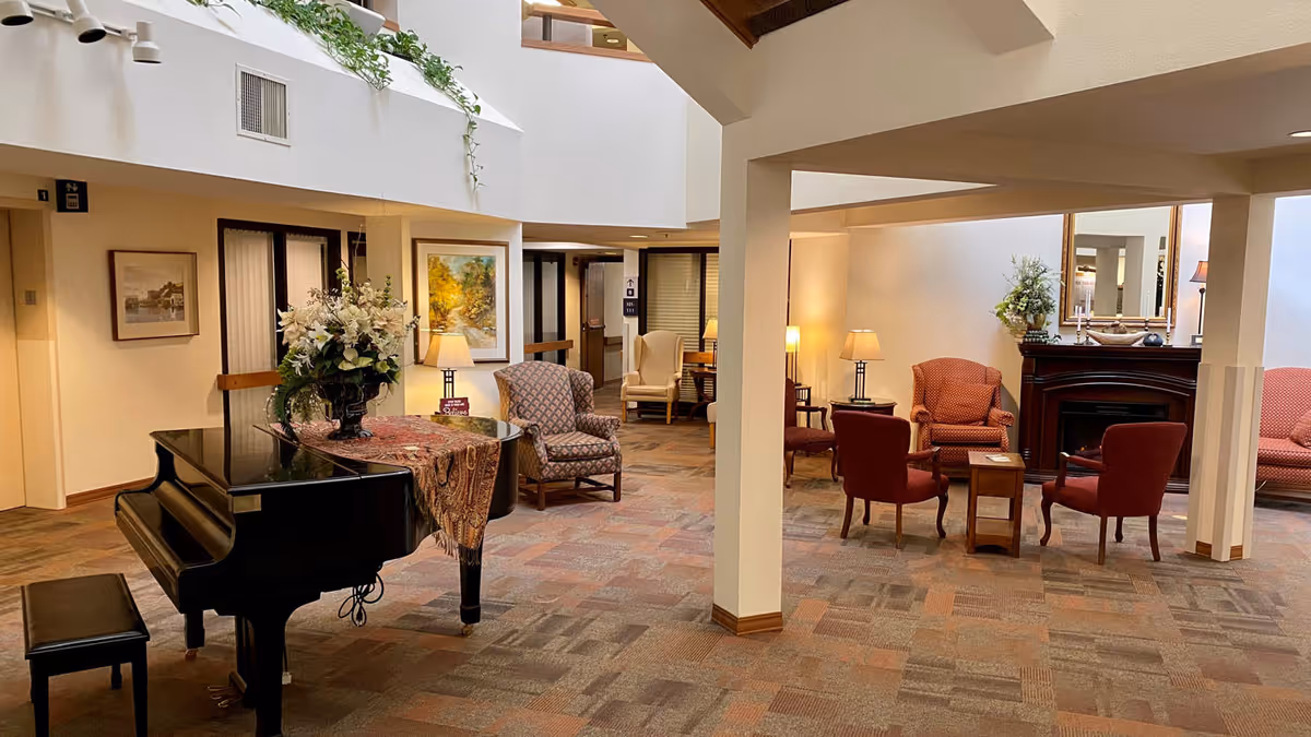 A bright communal lounge with a black grand piano, a floral centerpiece, multiple armchairs and a fireplace beneath an atrium skylight.