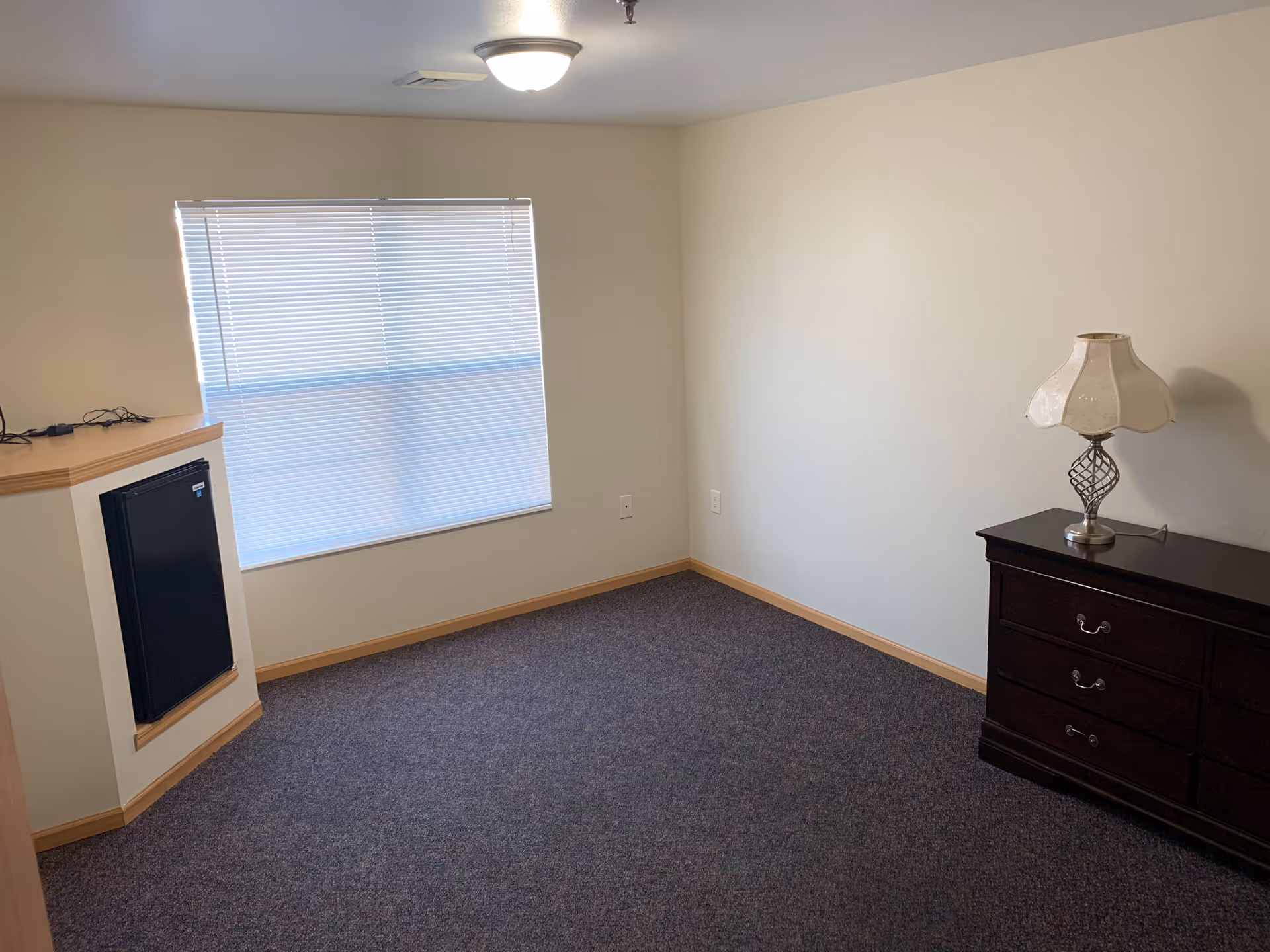 A small, empty room with beige walls and a dark carpeted floor. There is a window with closed blinds on one wall, a dark wooden dresser with a decorative lamp on top against another wall, and a small black mini-fridge built into a corner unit. The room is lit by a ceiling light fixture.