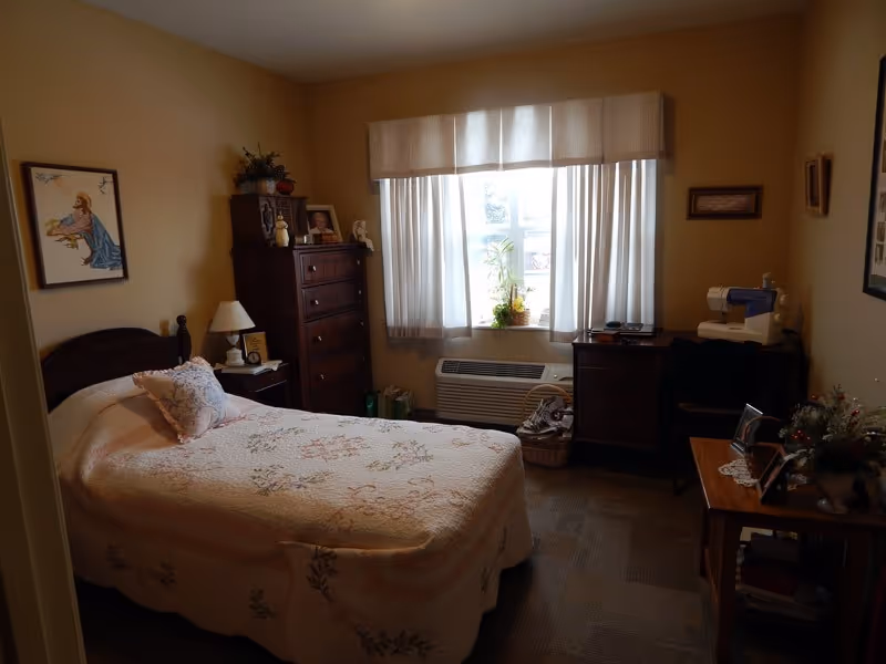 A cozy bedroom with a single bed covered in a floral quilt and a matching pillow. There is a wooden nightstand with a lamp and a framed picture next to the bed. A tall wooden dresser with decorative items and framed photos sits against the wall. A window with sheer curtains lets in natural light, and below it is a heating/cooling unit. On the right side, there is a desk with a sewing machine and a small table with flowers and framed photos. The walls are decorated with framed artwork and pictures.