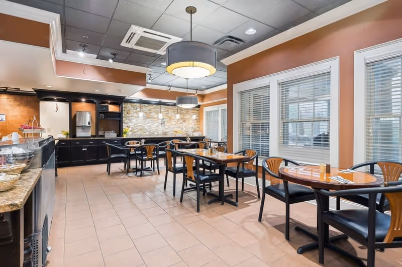 A dining room with several wooden tables and chairs arranged neatly. The room has large windows with white blinds, warm-colored walls, and modern ceiling lights. There is a counter area with a coffee machine and other items on the left side, and a stone accent wall in the background.