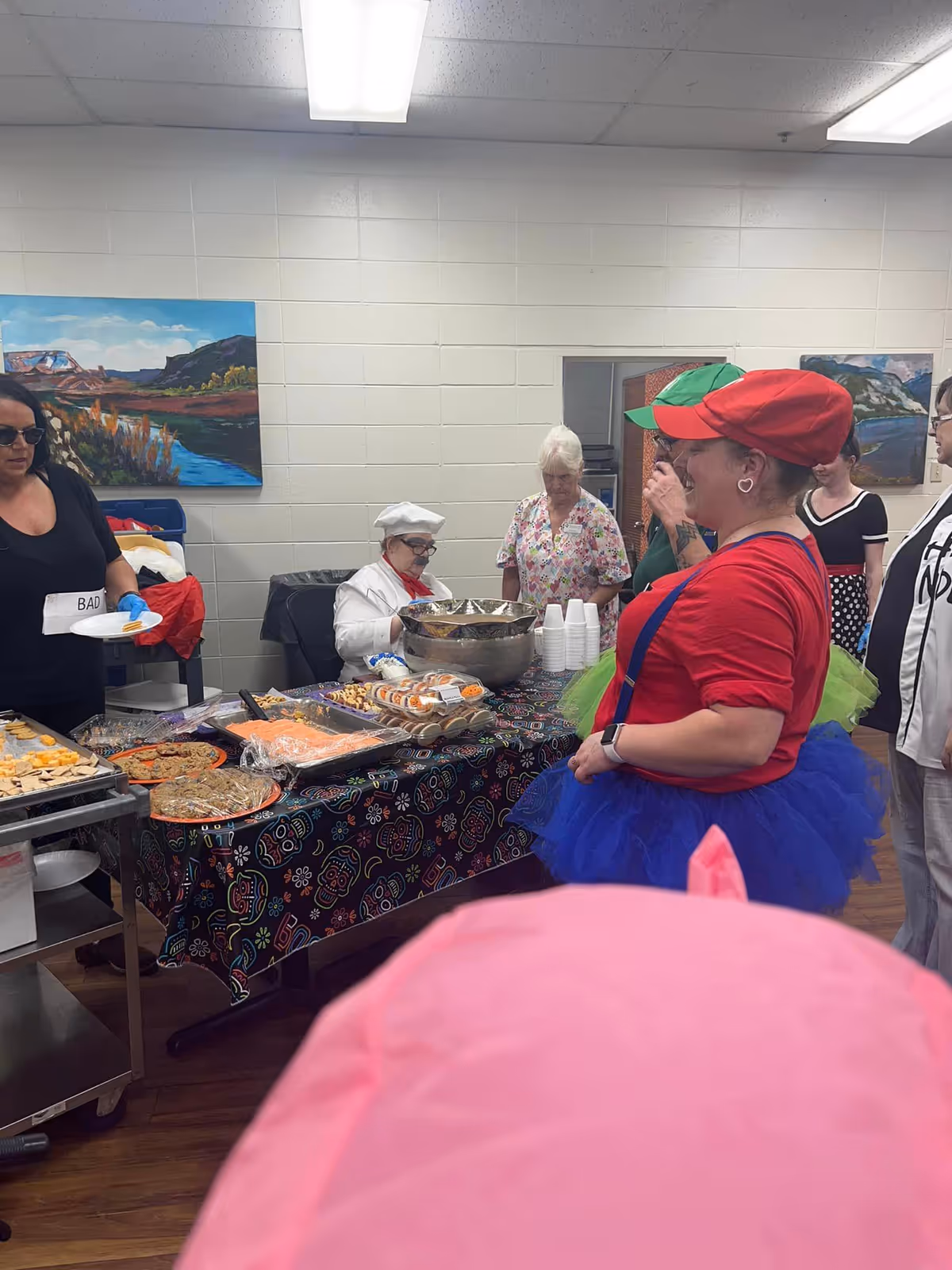 A group of people gathered around a table covered with a colorful tablecloth displaying various food items. One person is dressed as a chef with a white hat and coat, while another person wears a red shirt, blue tutu, and red cap. The setting appears to be an indoor room with white walls and landscape paintings hanging on the walls.