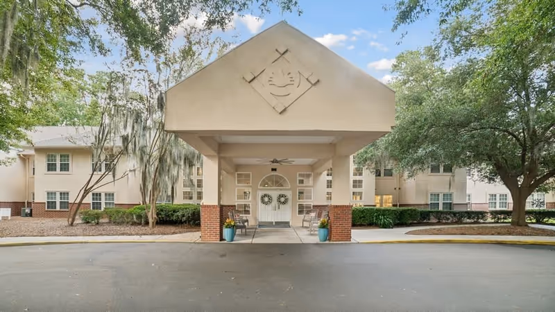 Front entrance of a senior living facility with a covered drop-off area supported by columns, two potted plants, and a double door decorated with wreaths. The building is surrounded by trees and landscaping under a partly cloudy sky.