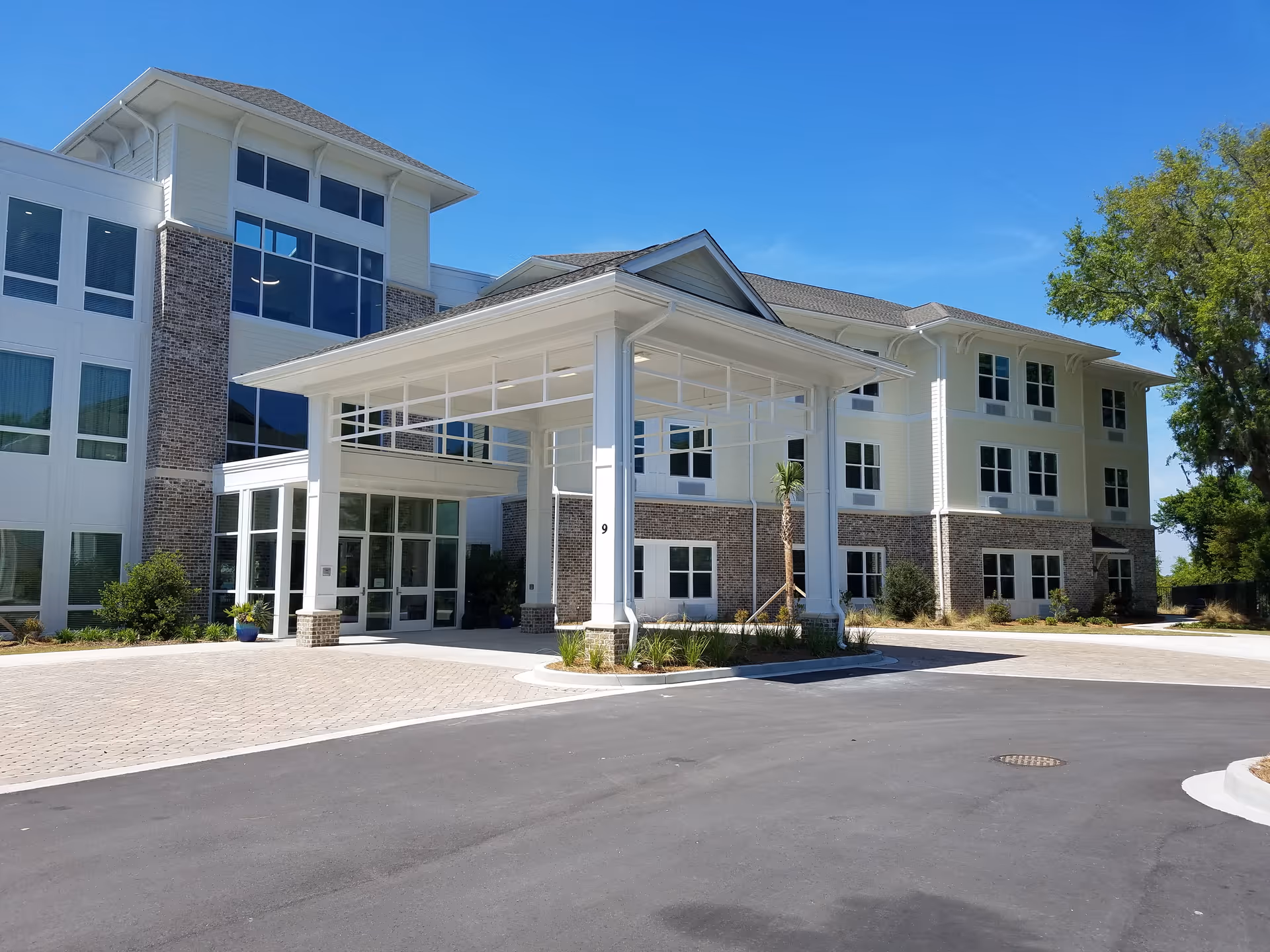 Exterior view of a modern three-story senior living facility building with a covered entrance, large windows, and a paved driveway under a clear blue sky.