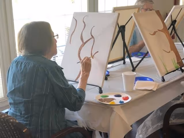 Two elderly women seated at a table indoors, each painting on a canvas set on easels. One woman is painting brown tree branches, and there are paint palettes and cups on the table. The room has large windows letting in natural light.