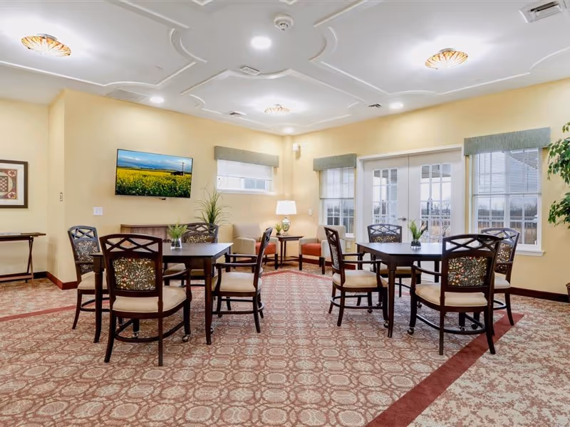 A bright and spacious common area with multiple round tables and chairs arranged on a patterned carpet. The walls are painted light yellow, and there are windows with green valances allowing natural light to enter. A flat-screen TV is mounted on one wall, and there are a couple of armchairs and a small table with a lamp in the corner. The ceiling features decorative molding and recessed lighting.