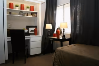 A small bedroom with a single bed covered in a brown bedspread, a black desk with a lamp and red decorative vases, a black chair, white built-in shelves with red candles and small plants, and a window with dark curtains.