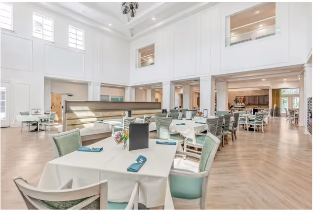 A bright, spacious dining room with multiple set tables and green-upholstered chairs beneath a high white coffered ceiling.