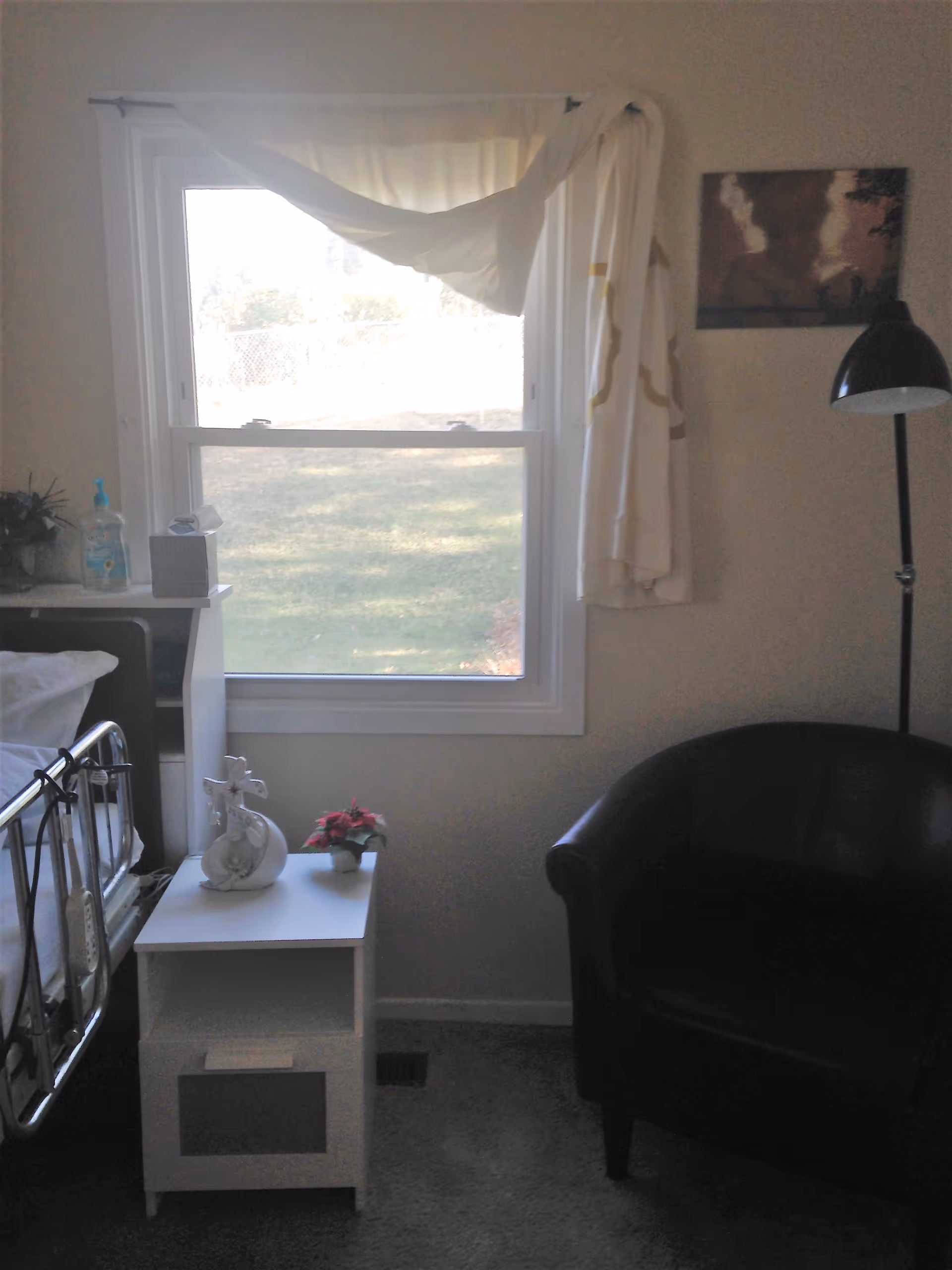 A small room with a window covered partially by white curtains. To the left is a hospital-style bed with a white nightstand next to it, holding a decorative cross, a small potted plant, a tissue box, and a bottle of hand sanitizer. To the right is a black armchair and a tall black floor lamp. A framed picture hangs on the wall above the chair.