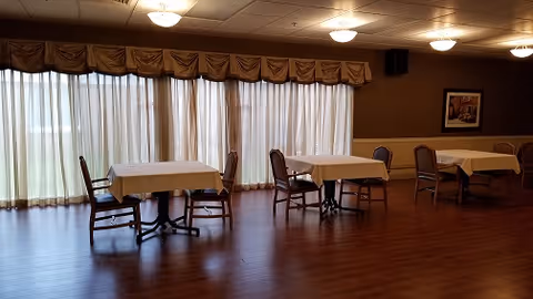 A dining room with wooden flooring and several tables covered with white tablecloths. Each table is accompanied by chairs, and large windows with sheer curtains and valances allow natural light to fill the room. The walls are decorated with framed artwork and the ceiling has multiple light fixtures.