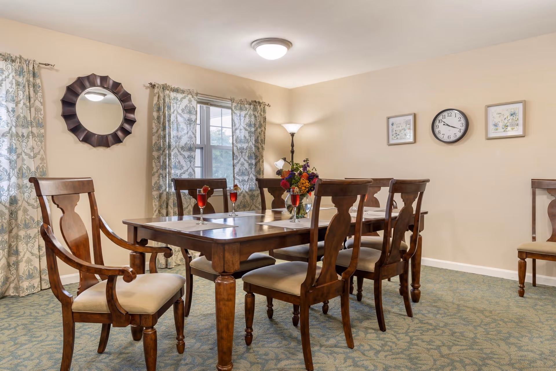 A dining room with a wooden dining table and six matching chairs with cushioned seats. The table is set with placemats, glasses filled with a red beverage, and a vase of colorful flowers. The room has patterned curtains on the windows, a round decorative mirror on one wall, a floor lamp in the corner, and a clock and two framed pictures on another wall. The carpet has a subtle leaf pattern.